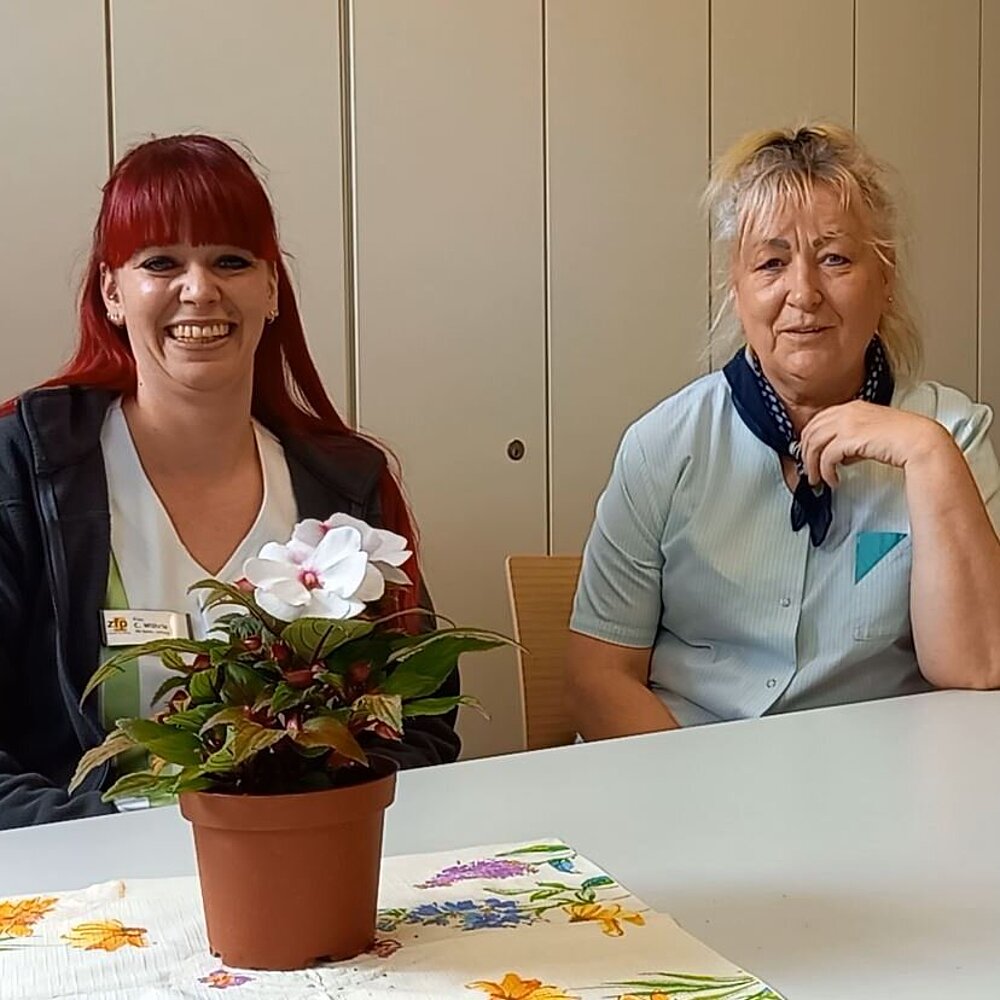 Zwei Frauen, eine jüngeren Alters mit roten langen Haaren, die andere blond und mittleren Alters, sitzen lachend an einem Tisch, auf welchem kleine Blumengestecke stehen.