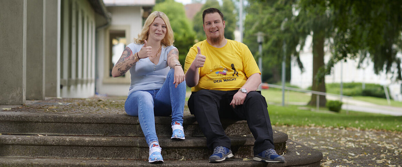 Eine junge Frau mit langen blonden Haaren und ein junger Mann im gelben T-Shirt sitzen auf Stufen vor einem Gebäude. 