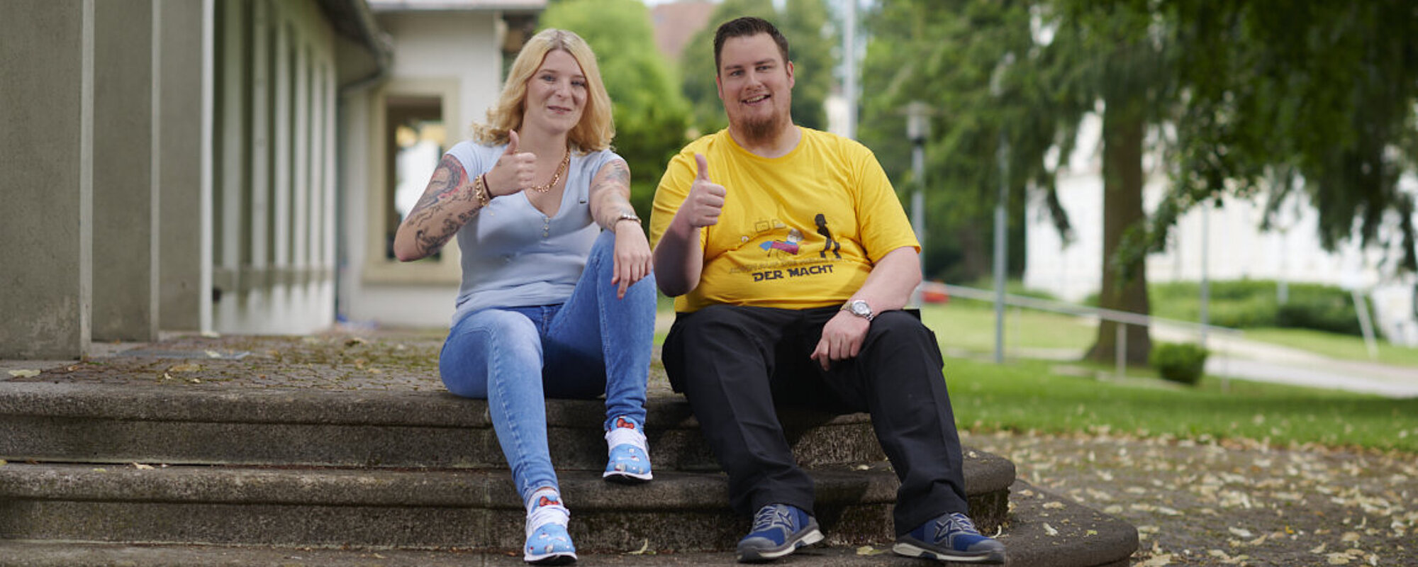 Eine junge Frau mit langen blonden Haaren und ein junger Mann im gelben T-Shirt sitzen auf Stufen vor einem Gebäude. 