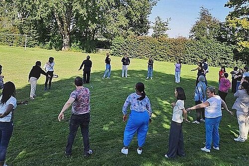 Eine Gruppe Menschen steht in einem großen Kreis auf einer Wiese, einige Personen schwingen die Arme. 