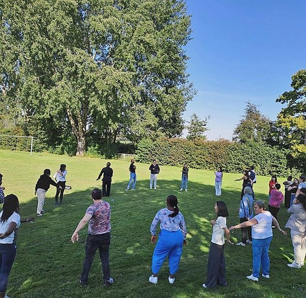 Eine Gruppe Menschen steht in einem großen Kreis auf einer Wiese, einige Personen schwingen die Arme. 