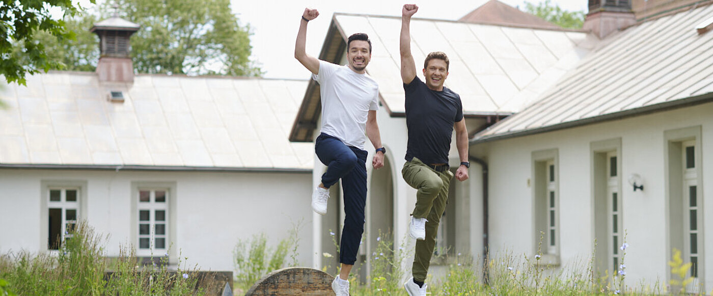 Zwei junge Männer springen dynamisch in die Höhe und strecken jeweils einen Arm nach oben. 