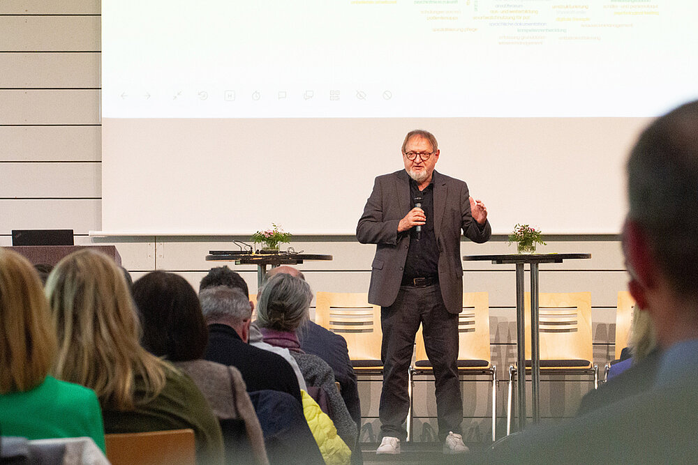 Ein Mann mit Brille hält eine Präsentation auf einer Tagung. Er spricht vor einem Publikum und verwendet eine Präsentationsfolie.