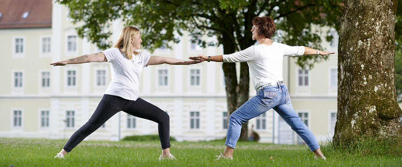 Eine junge Frau und eine ältere Frau stehen sich in einer Yogahaltung gegenüber. 