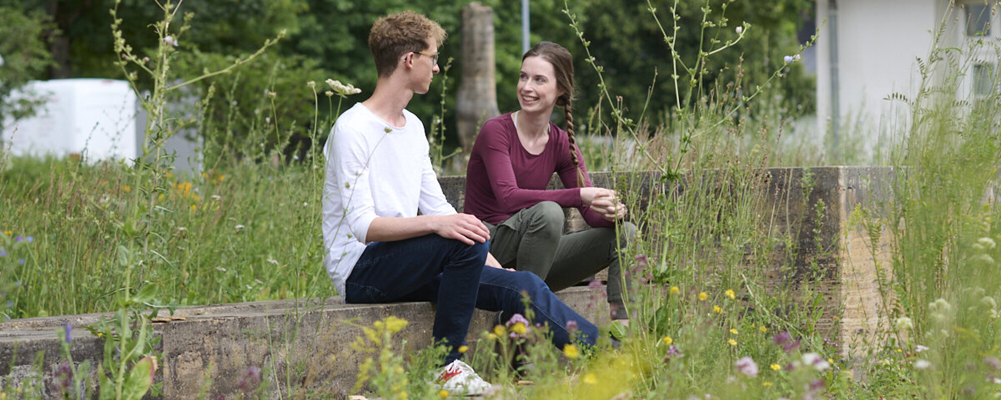 Ein junger Man und eine junge Frau sitzen sich auf einem Steinblock gegenüber und sind in ein Gespräch vertieft. 