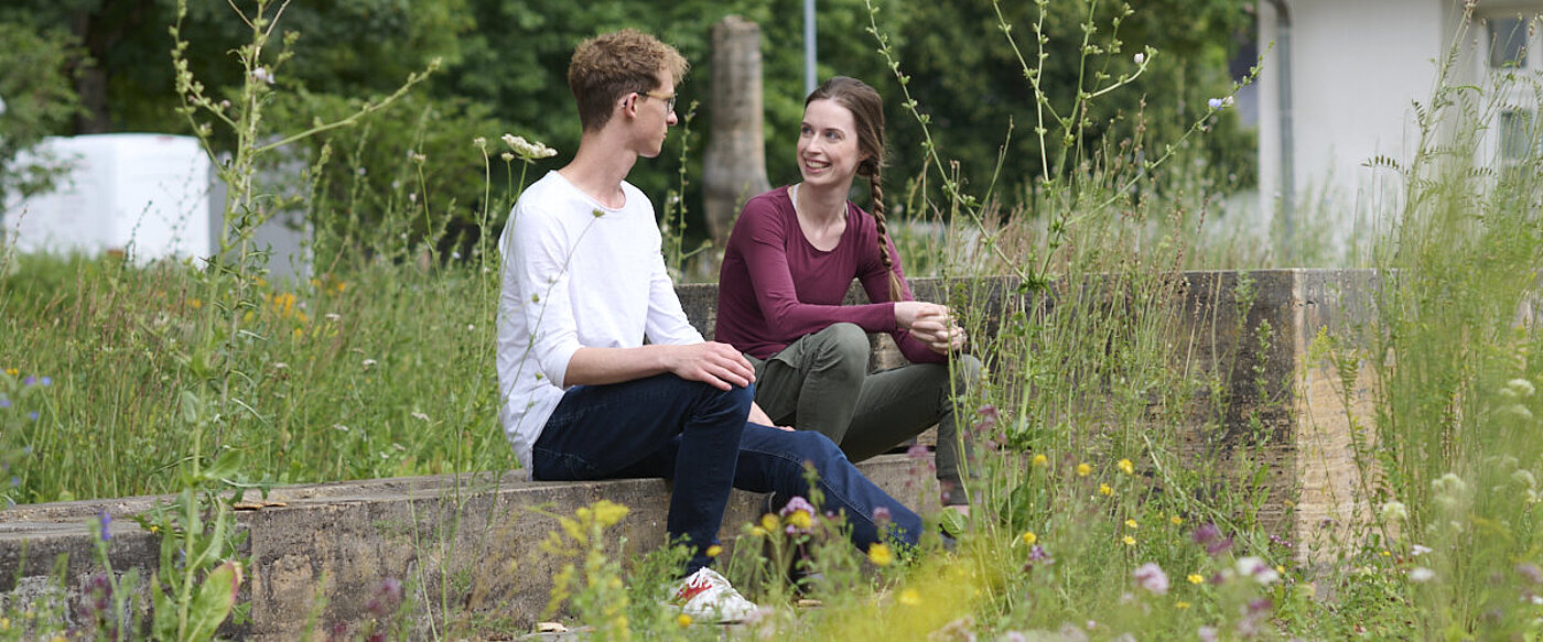 Ein junger Man und eine junge Frau sitzen sich auf einem Steinblock gegenüber und sind in ein Gespräch vertieft. 