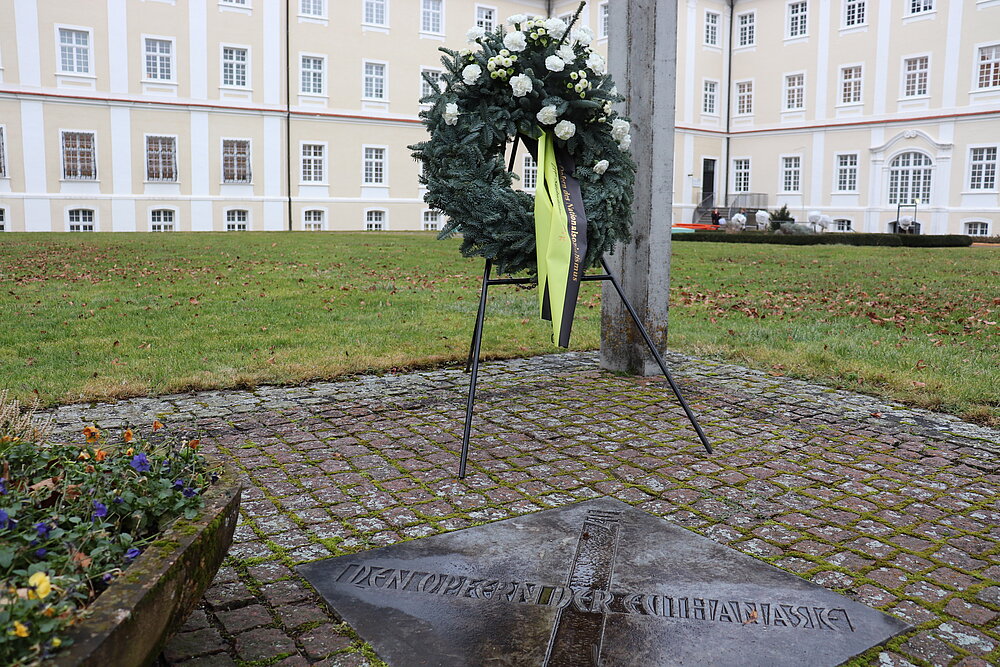 Am Mahnmal Offenes Haus ist ein geschmückter Kranz aufgestellt. Im Hintergrund ist das Kloster Bad Schussenried zu sehen.