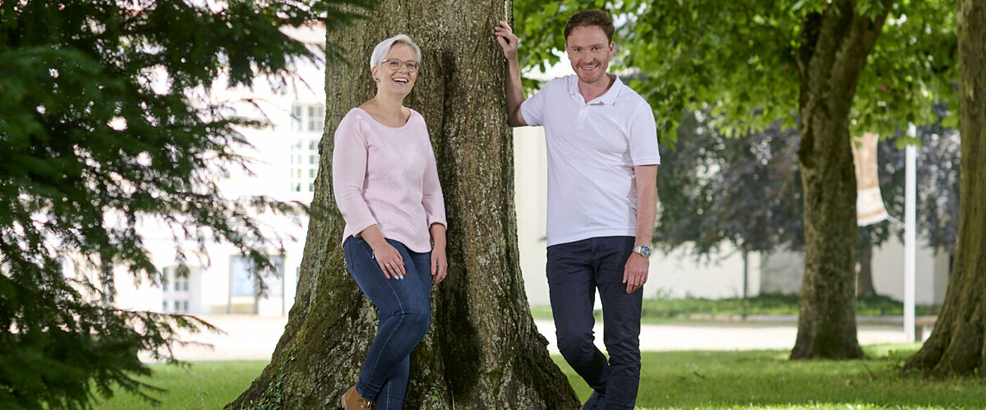 Eine junge Frau und ein Mann im mittleren Alter stehen vor einem großen Baum. 