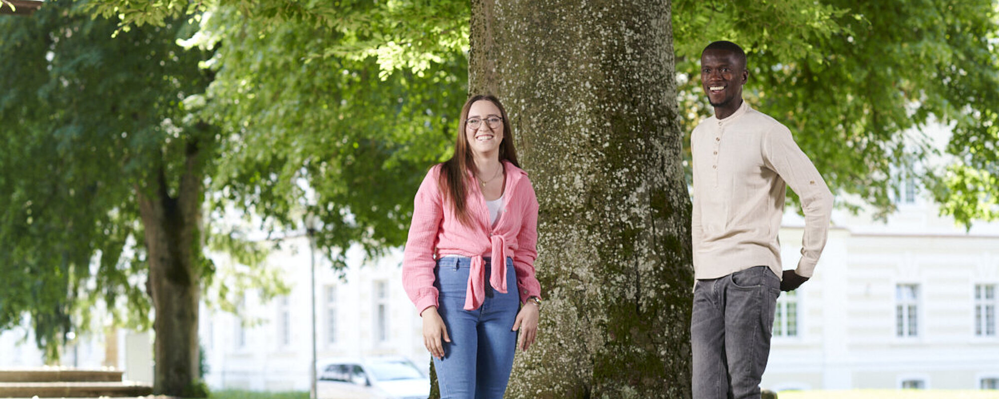 Eine junge Frau und ein junger Mann stehen vor einem großen Baum und recken die Arme in die Höhe. 