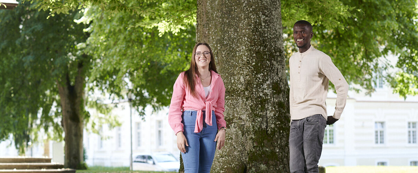 Eine junge Frau und ein junger Mann stehen vor einem großen Baum und recken die Arme in die Höhe. 