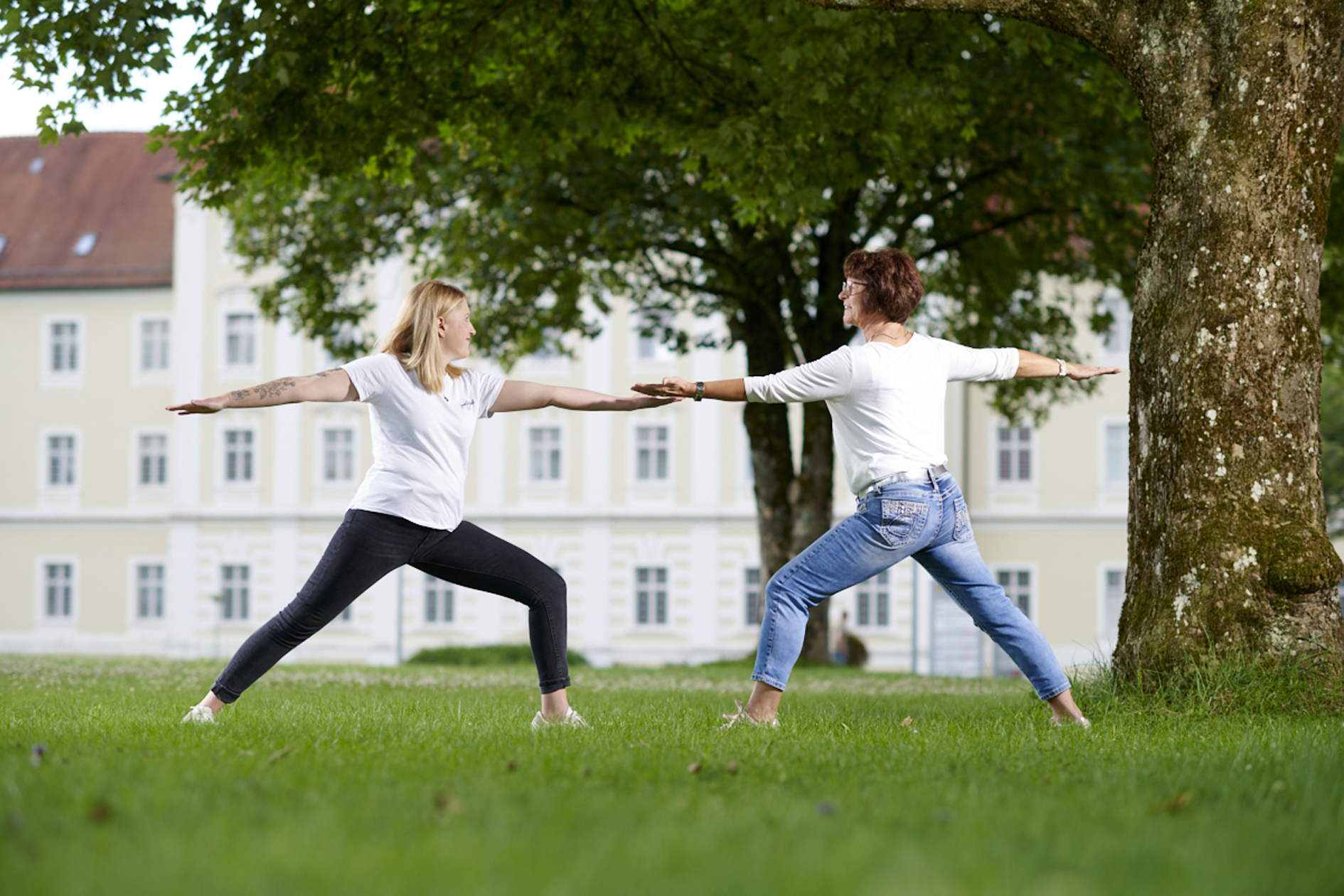 Eine junge Frau und eine ältere Frau stehen sich in einer Yogahaltung gegenüber. 